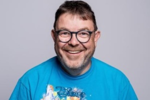 Ein Mann mit einem blauen Shirt und einer Brille grinst breit in die Kamera.